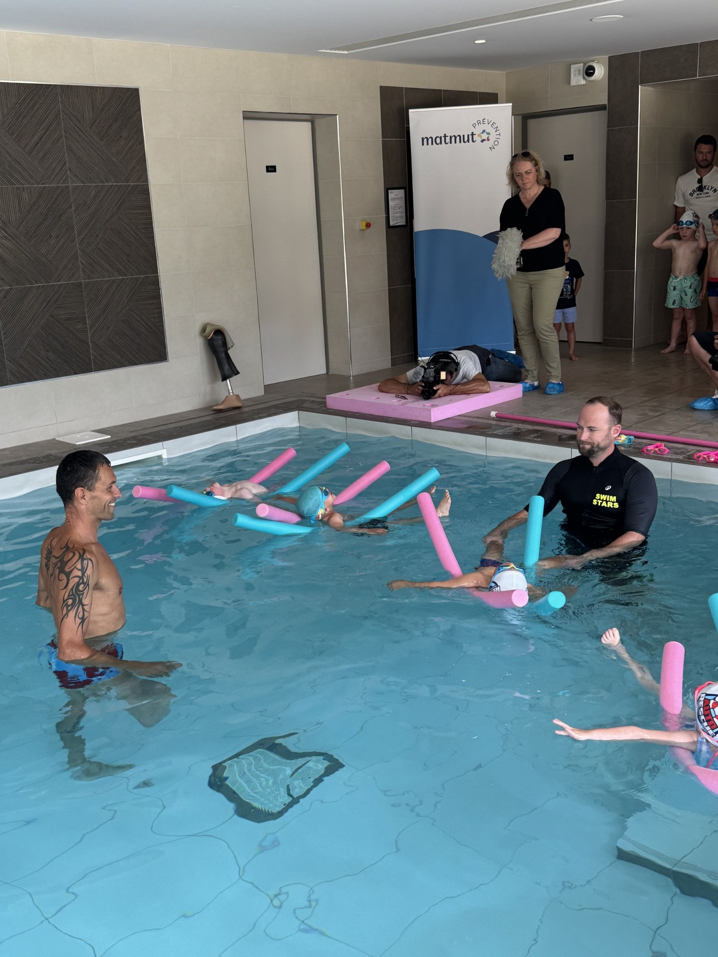 Alexis Hanquinquant dans le bassin Swim Stars avec un coach et des enfants, banderole Matmut Prévention en arrière-plan